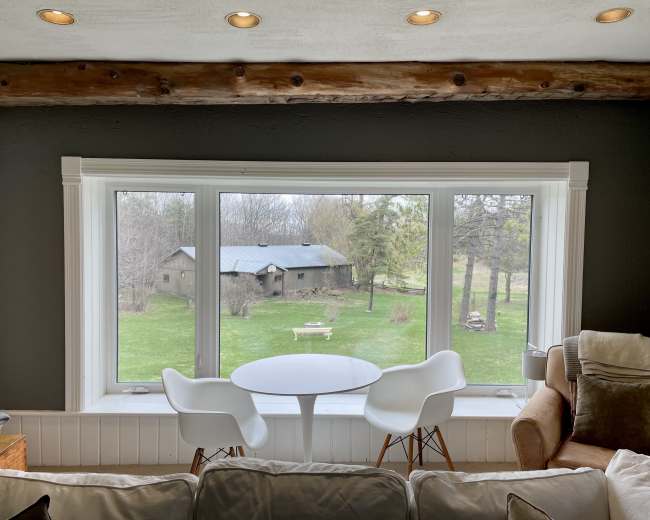 A window view featuring a green lawn, a small barn, and two white chairs with a round table in front of the window.