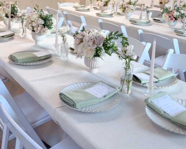 A long table is set for a formal dinner, complete with white chairs, elegant tableware, and floral centerpieces.