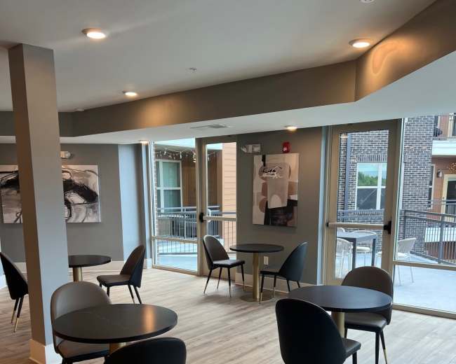 The image shows a modern lounge area with several round tables and black chairs, featuring large windows that provide natural light.