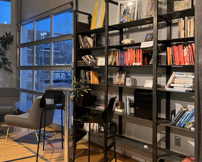 A modern indoor space with a metal bookshelf filled with books, a small table, and chairs, illuminated by soft evening light coming through large windows.