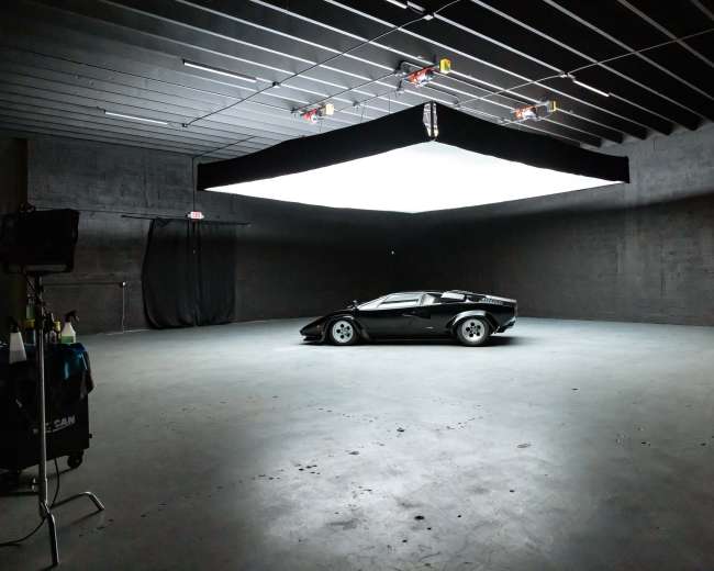 A black sports car is positioned in a dimly lit studio with a large overhead light source illuminating the scene.