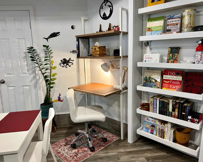 The image shows a compact workspace with a wooden desk, a swivel chair, a plant, and shelves filled with books and decorative items.