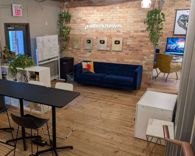 The image shows a modern workspace featuring a seating area with a blue couch, a dining table with chairs, and an exposed brick wall displaying framed plaques.