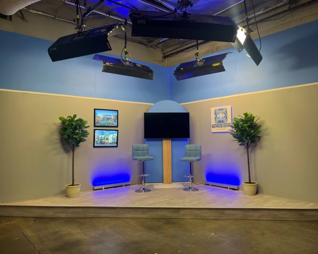 The image shows a television studio set featuring two blue bar stools, a central television screen, and decorative plants against a light blue and gray background.
