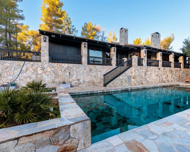 The image shows a stone-lined swimming pool in front of a rustic, two-story house surrounded by trees.