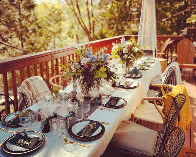 A long dining table set with elegant plates and glasses is adorned with floral centerpieces on a tree-lined deck.