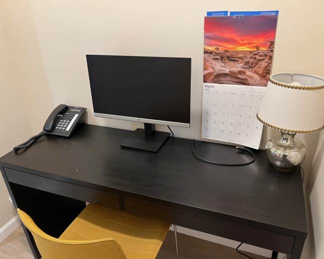 A black desk with a computer monitor, a telephone, a lamp, and a calendar is set against a beige wall.