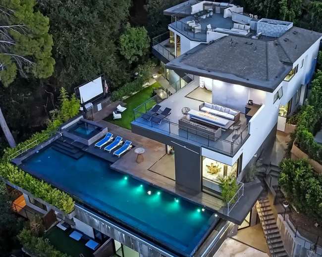 The image shows a modern multi-story house with a large rooftop terrace and an infinity pool surrounded by landscaping and lounge chairs.