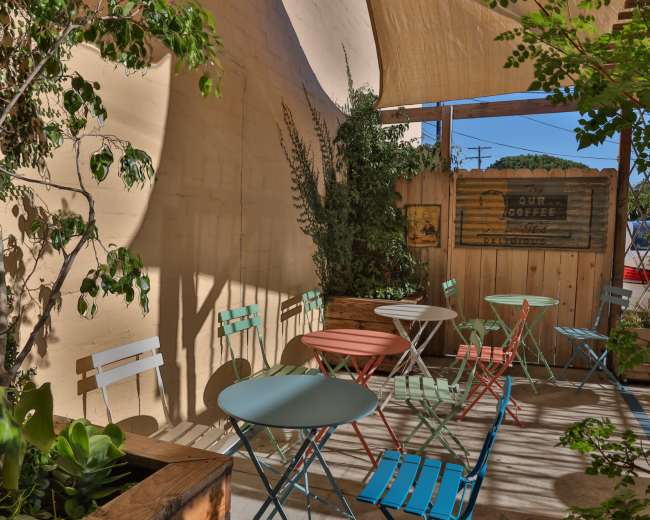 The image shows a small outdoor café area with colorful metal chairs and tables surrounded by greenery and wooden fencing.