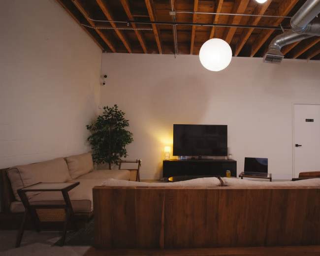 The image shows a modern living room with a sectional couch, a television on a media console, and a potted plant, all beneath hanging light fixtures and exposed wooden beams.