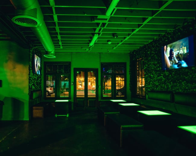The image shows a dimly lit lounge with green lighting, featuring low benches and two televisions on the walls.