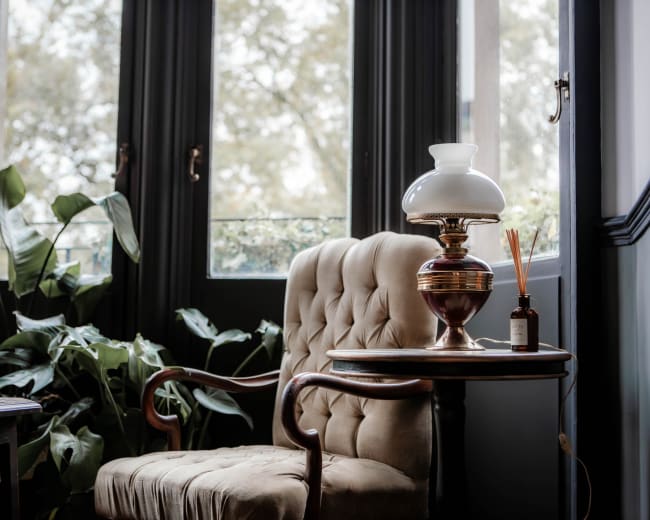 A tufted armchair is positioned next to a small table with a vintage lamp and a decorative bottle, surrounded by large plants and sunlight filtering through dark windows.