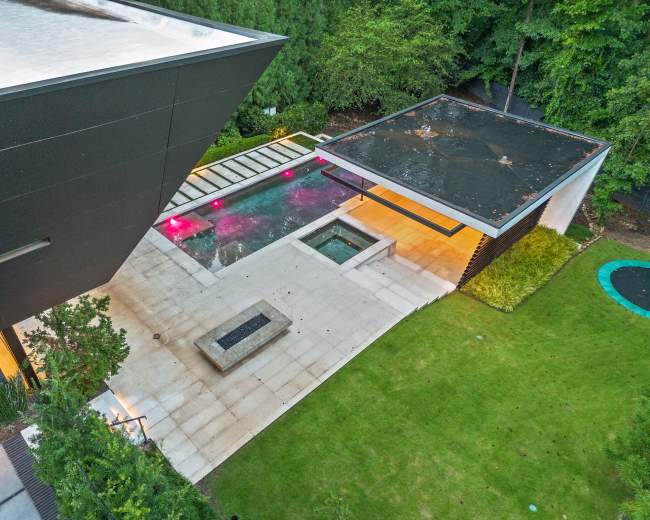 The image shows a modern outdoor space featuring a swimming pool, a hot tub, a covered patio area, and a green lawn surrounded by trees.