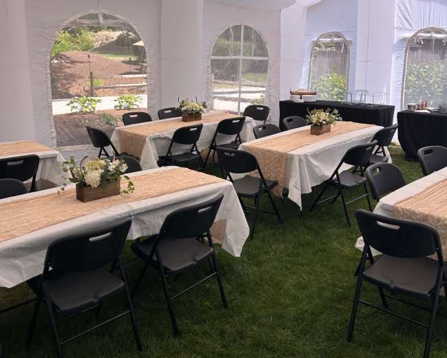 The image shows a large white tent set up with several tables decorated with table runners and floral centerpieces, all arranged on a grassy area.