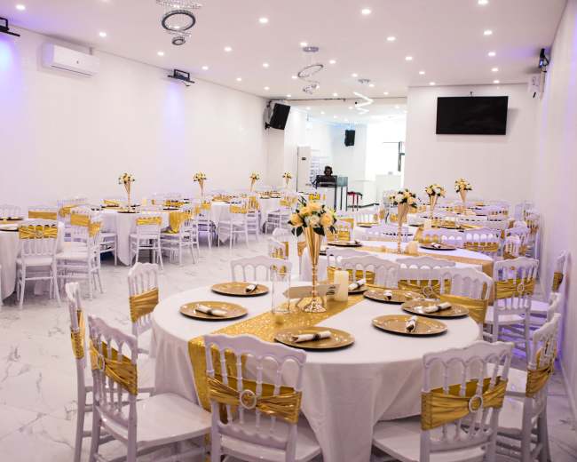 A banquet hall is set up with round tables covered in white tablecloths, gold accents, and floral centerpieces, ready for an event.
