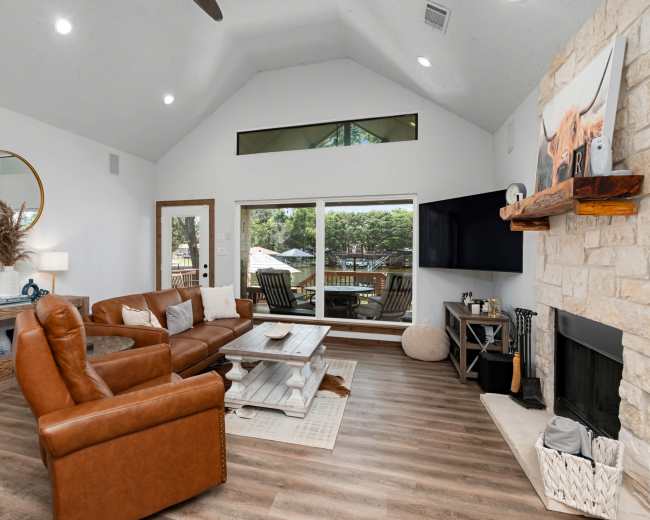 The living room features a brown leather sectional sofa, a wooden coffee table, and large windows that overlook an outdoor seating area.