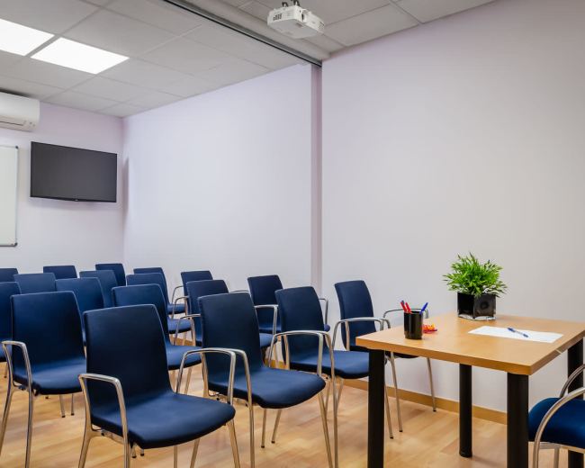 The image shows a conference room with several blue chairs arranged in rows facing a small table and a projector screen.