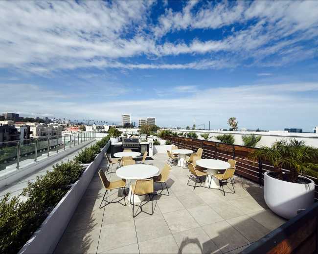 The image shows a rooftop terrace featuring several round tables and chairs, with a view of buildings and palm trees under a partly cloudy sky.