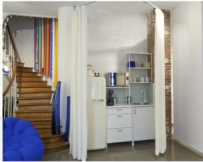 The image shows a small kitchen area enclosed by curtains, adjacent to a wooden staircase and featuring a mix of modern and rustic elements.