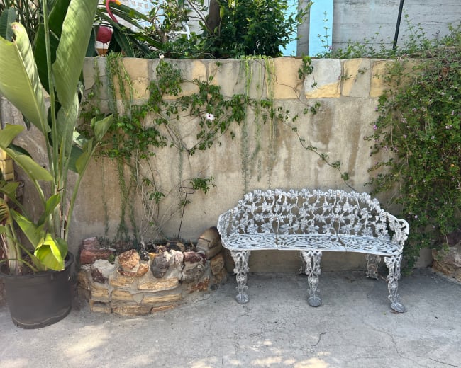 A white cast iron bench with intricate designs is set against a weathered stone wall covered with vines and plants.