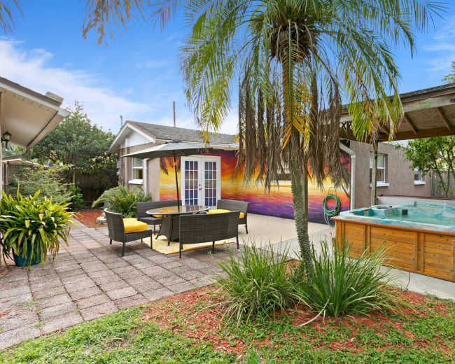 A backyard patio with a dining table and chairs, a hot tub, a colorful mural on the wall, and surrounding greenery.
