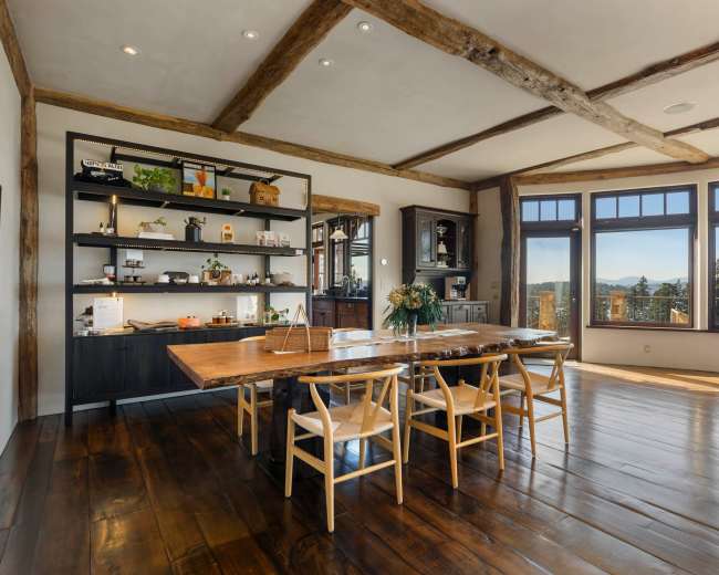 A spacious dining area features a large wooden table surrounded by chairs and a shelf displaying various items, with large windows offering a view of the outdoors.