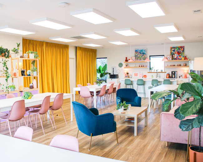 A modern office space with multiple work tables, colorful chairs, a kitchenette area, and large windows with yellow curtains.