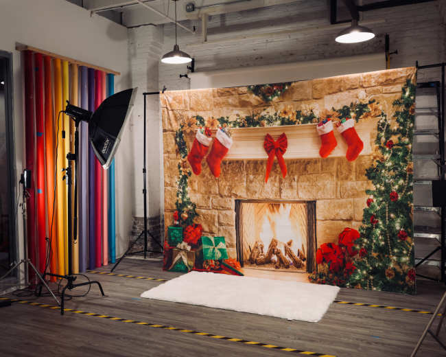 The image shows a holiday-themed photography backdrop featuring a decorated fireplace with stockings and gifts, set in a studio with colorful vertical stripes in the background.