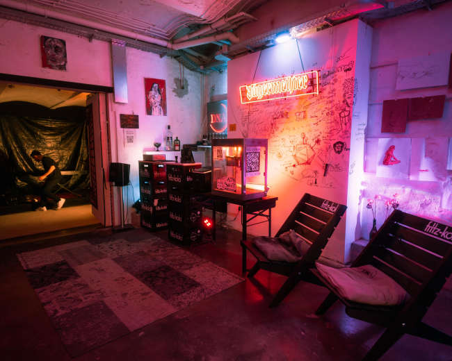 The image shows a creatively designed room featuring colorful neon lights, a vintage popcorn machine, and artistic wall decorations, with a person sitting in the background.