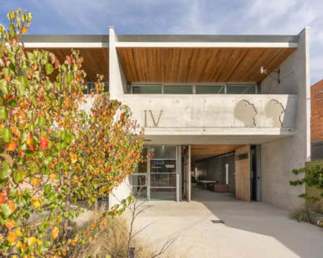 The image shows a modern building with a concrete facade and large glass entrance, surrounded by landscaping featuring colorful foliage.