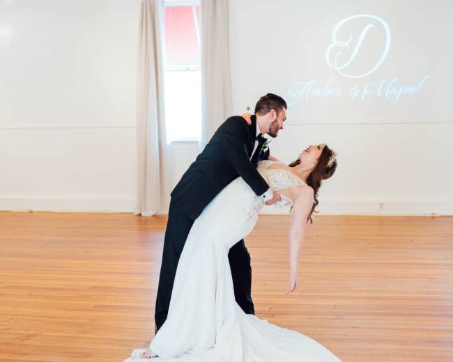 A bride and groom share a dance in a well-lit room with wooden floors and a chandelier overhead.