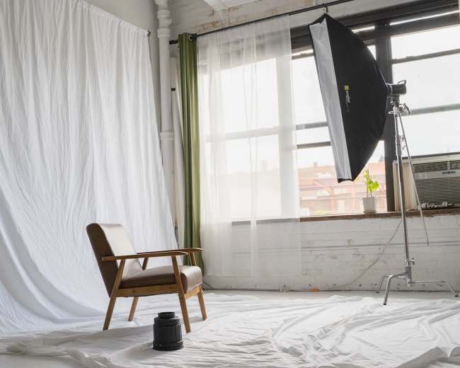 A wooden chair is positioned in a brightly lit room with a white backdrop and soft fabric curtains.