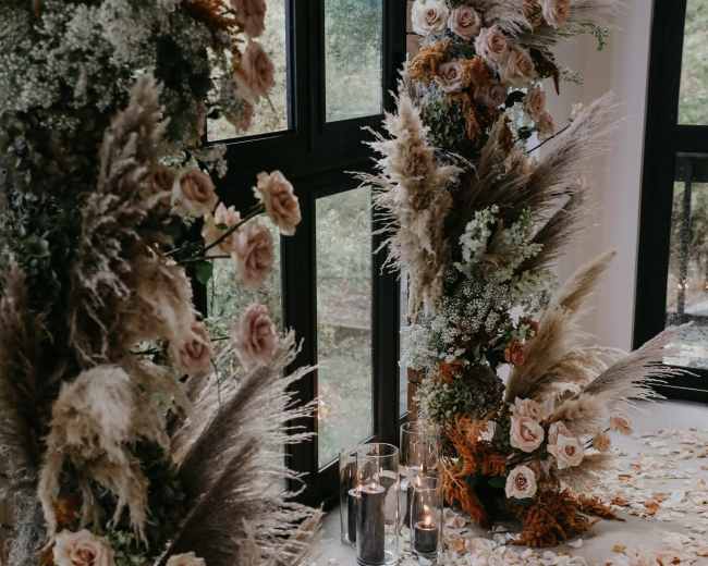 The image features two floral arrangements made of roses and pampas grass beside a carpet of rose petals and lit candles on the floor.