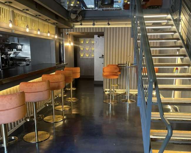 The image shows a modern interior of a restaurant with orange bar stools along a bar, a staircase leading to an upper level, and soft lighting.
