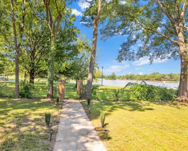 A stone pathway leads through a green yard lined with trees to a calm lake in the background.