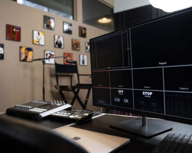 A computer monitor displaying video production controls is positioned on a desk in a studio adorned with framed artwork.
