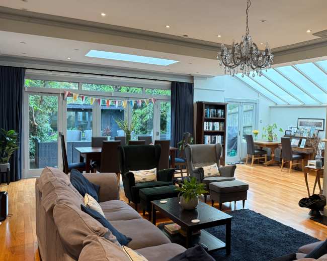 The image shows a spacious living room with modern furnishings, a fireplace, a large skylight, and a dining area visible through glass doors.