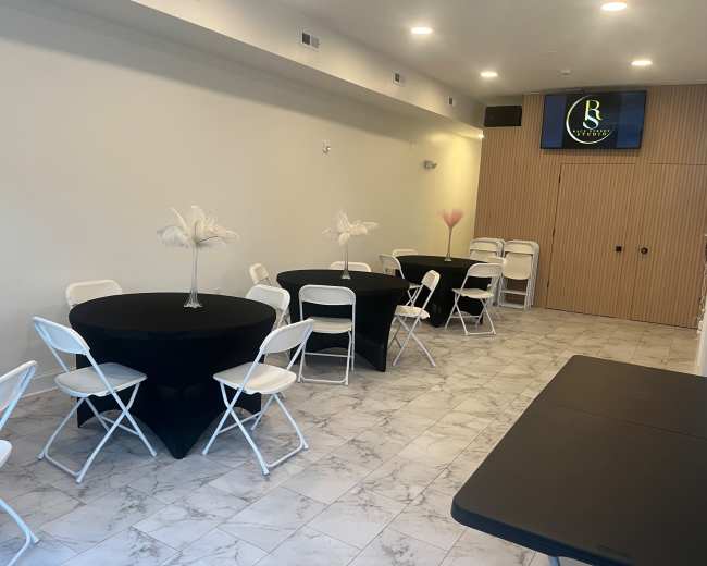 The image shows an interior space with several round tables covered in black tablecloths, white folding chairs, and a screen displaying a logo on the wall.