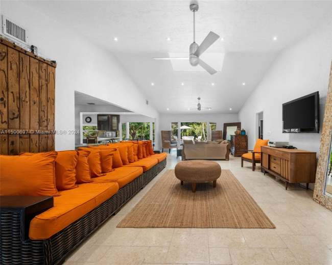 The image shows a spacious interior living area featuring a large orange-cushioned sofa, a central round coffee table, a flat-screen TV on the wall, and a mirror reflecting part of the room.