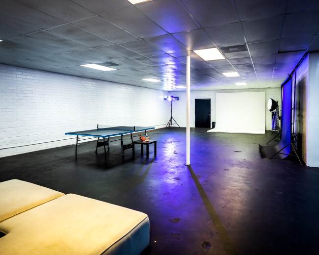 The image shows a spacious, empty room with a table tennis table, a small table, and various lighting equipment in the corners.