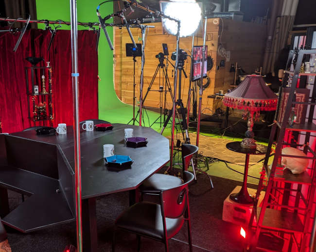 The image shows a film set featuring a round table surrounded by chairs, with a backdrop of red curtains and various lighting and camera equipment positioned around the scene.