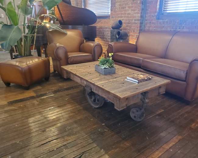 The image shows a living room with a leather sofa, a matching armchair, and a wooden coffee table with wheels, all arranged on a wooden floor.