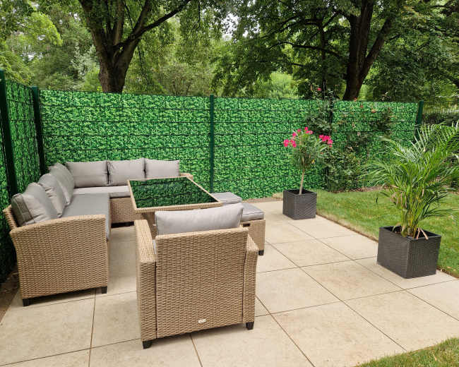 The image shows a landscaped outdoor seating area with a wicker sofa set, glass coffee table, and potted plants surrounded by a green privacy fence.