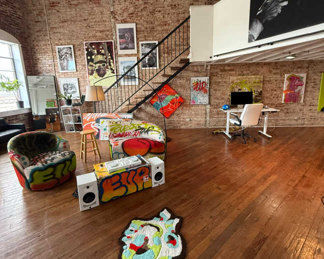 The image shows a spacious, industrial-style office with exposed brick walls, a staircase, vibrant graffiti-covered furniture, and a desk area equipped with a computer.