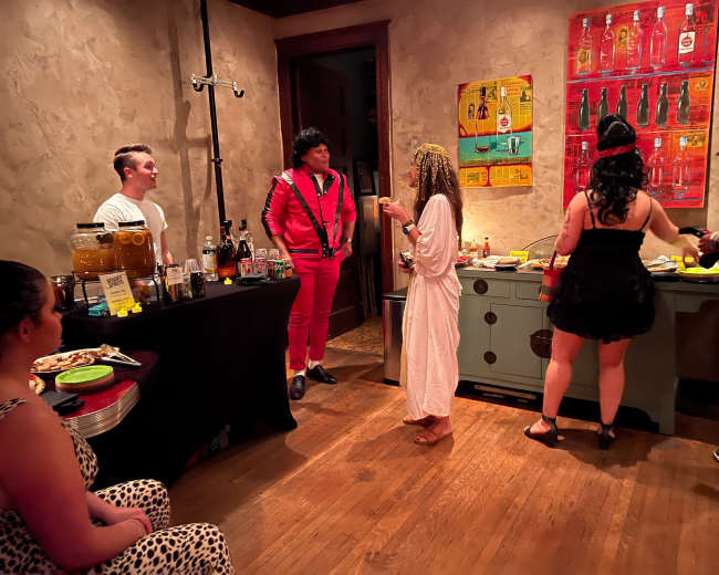 A small gathering is taking place in a room with a mix of people socializing around a bar setup, with drinks and snacks on display.