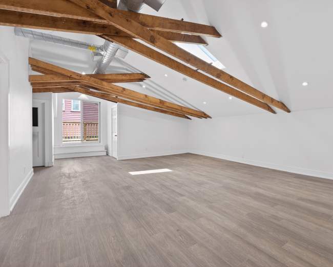 The image shows an empty room with wooden beams on the ceiling and large windows allowing natural light to enter.