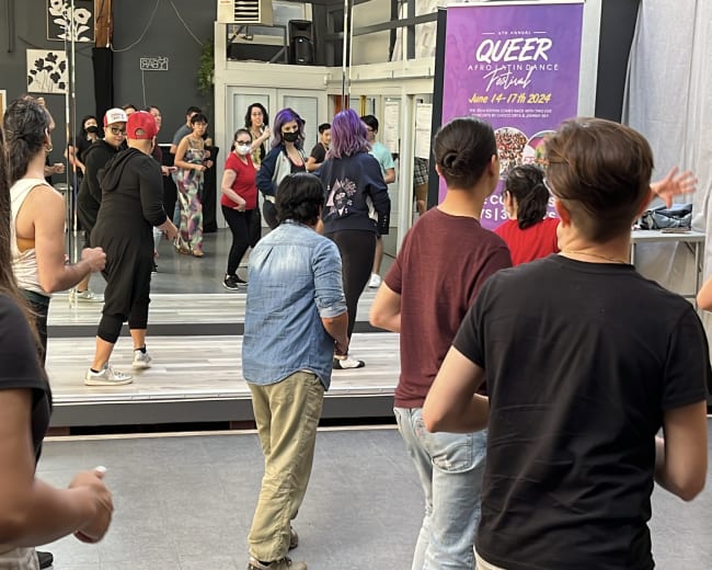 A group of individuals participates in a dance class at a studio, with mirrors reflecting their movements and a promotional poster for a queer event visible on the wall.