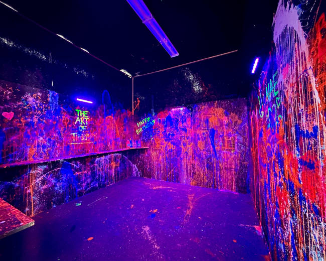 The image shows a dimly lit room with vibrant splashes of paint on the walls and shelves, illuminated by blacklight.