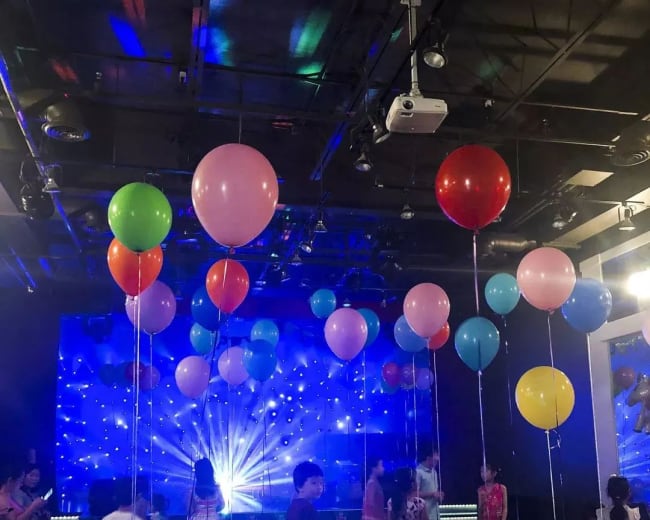 Children are playing with colorful balloons in a brightly lit party room with a light display.