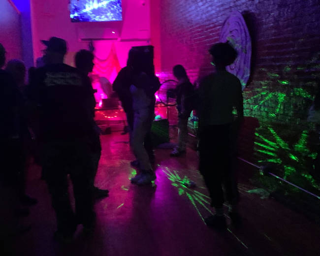 A group of people dance under colorful lights in a dimly lit venue with a stage in the background.